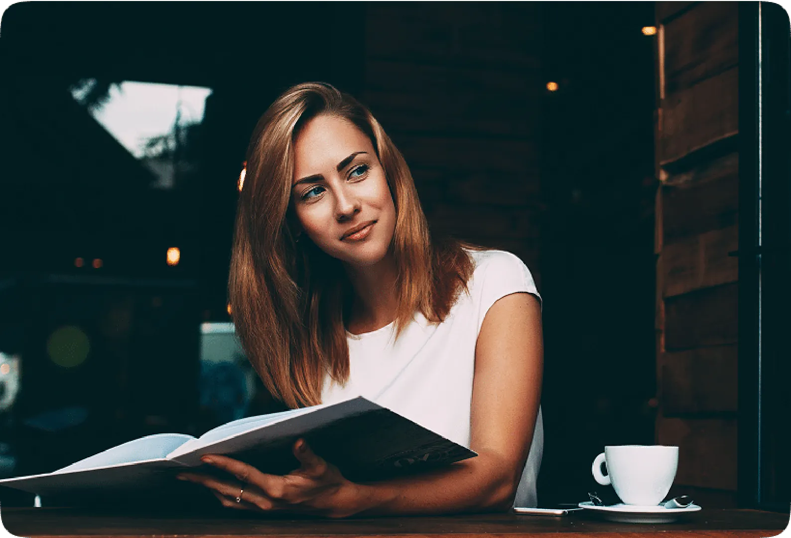A woman in a white shirt sits at a wooden table, reading a magazine with a coffee cup beside her.
