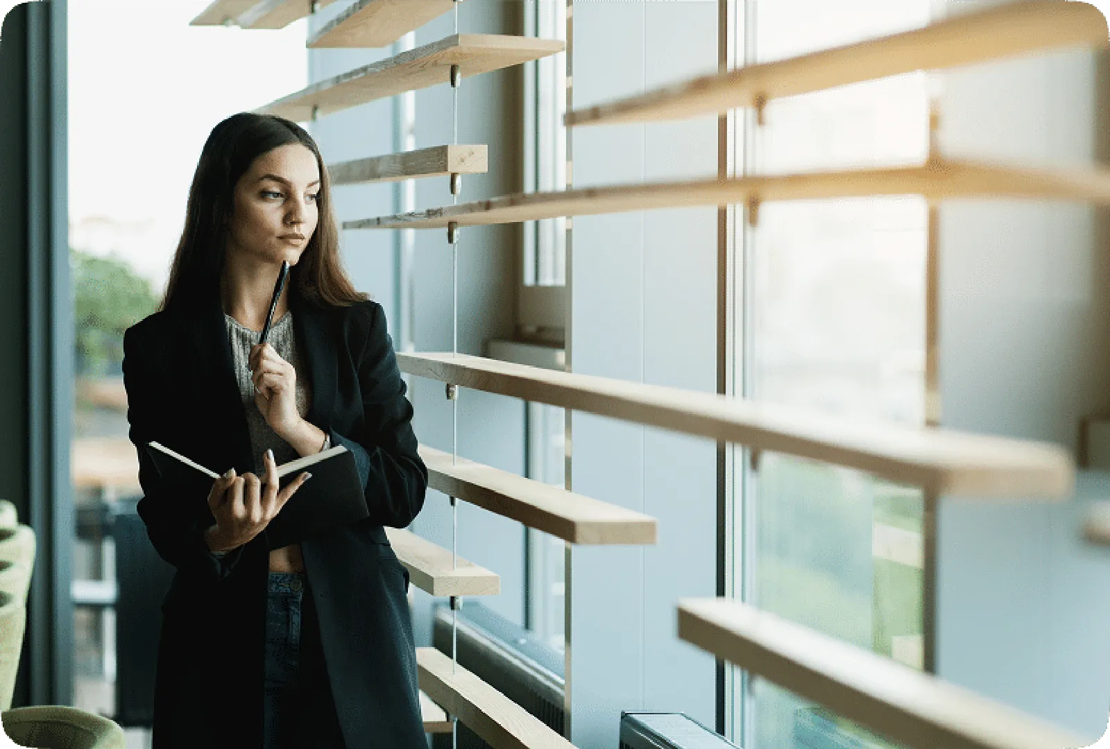 A woman wearing a dark dress, holding a book and pen, gazing thoughtfully by a window.