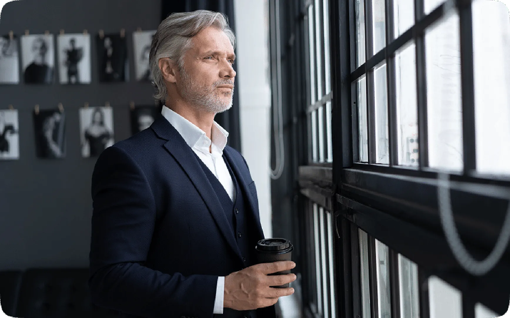 Man in a black suit by the window, gazing thoughtfully and holding a cup of coffee.