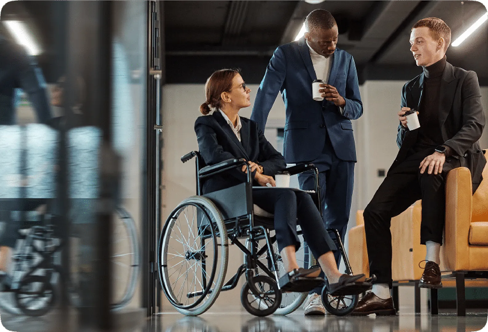 A professional group discussion featuring a woman in a wheelchair and two men with coffee cups.