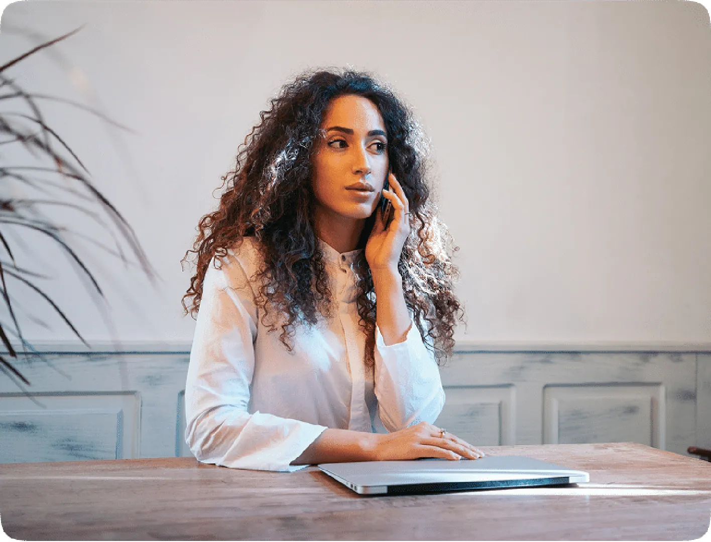 Woman on the phone with dark curly hair in a white shirt, sitting with a laptop.