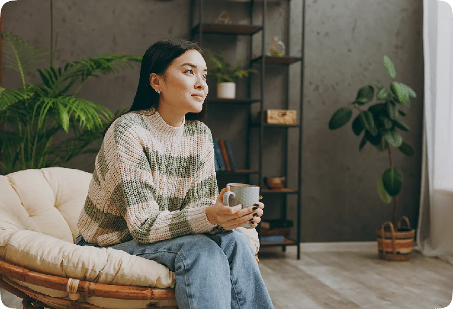 A smiley woman in a warm sweater and light jeans sitting on a cozy sofa holding a cup.