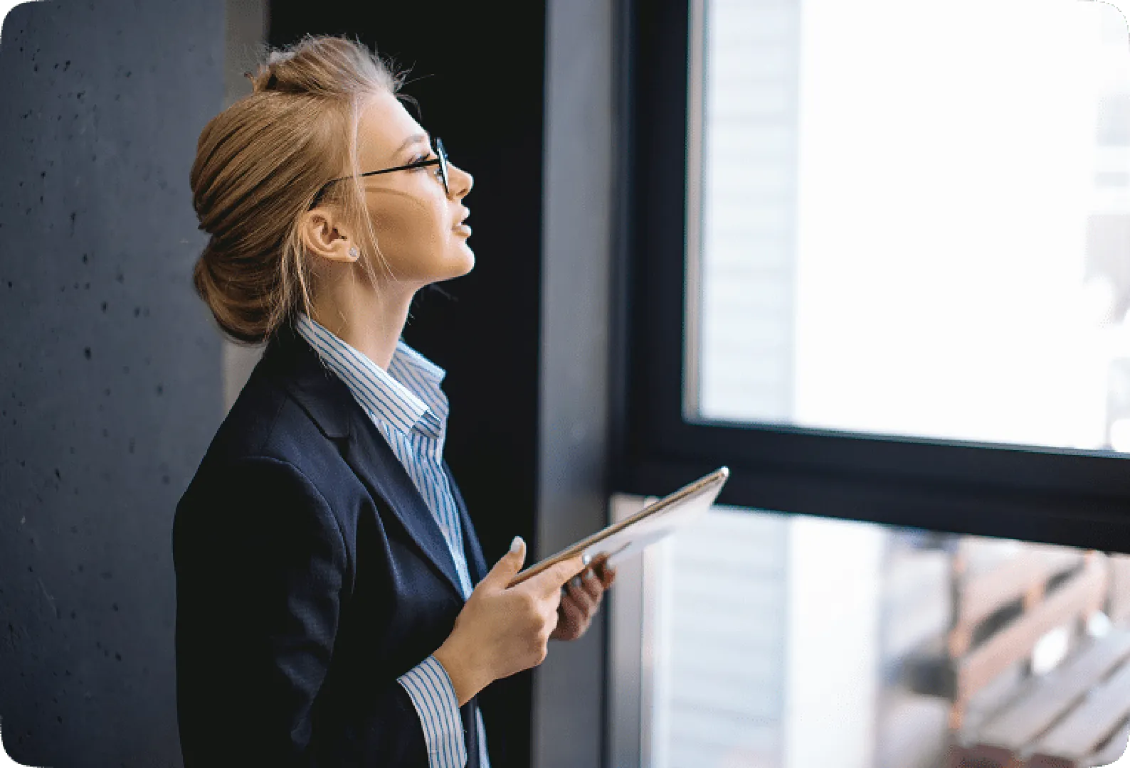 A smiley woman in a black suit wearing glasses, holding a laptop and looking outside by the window.