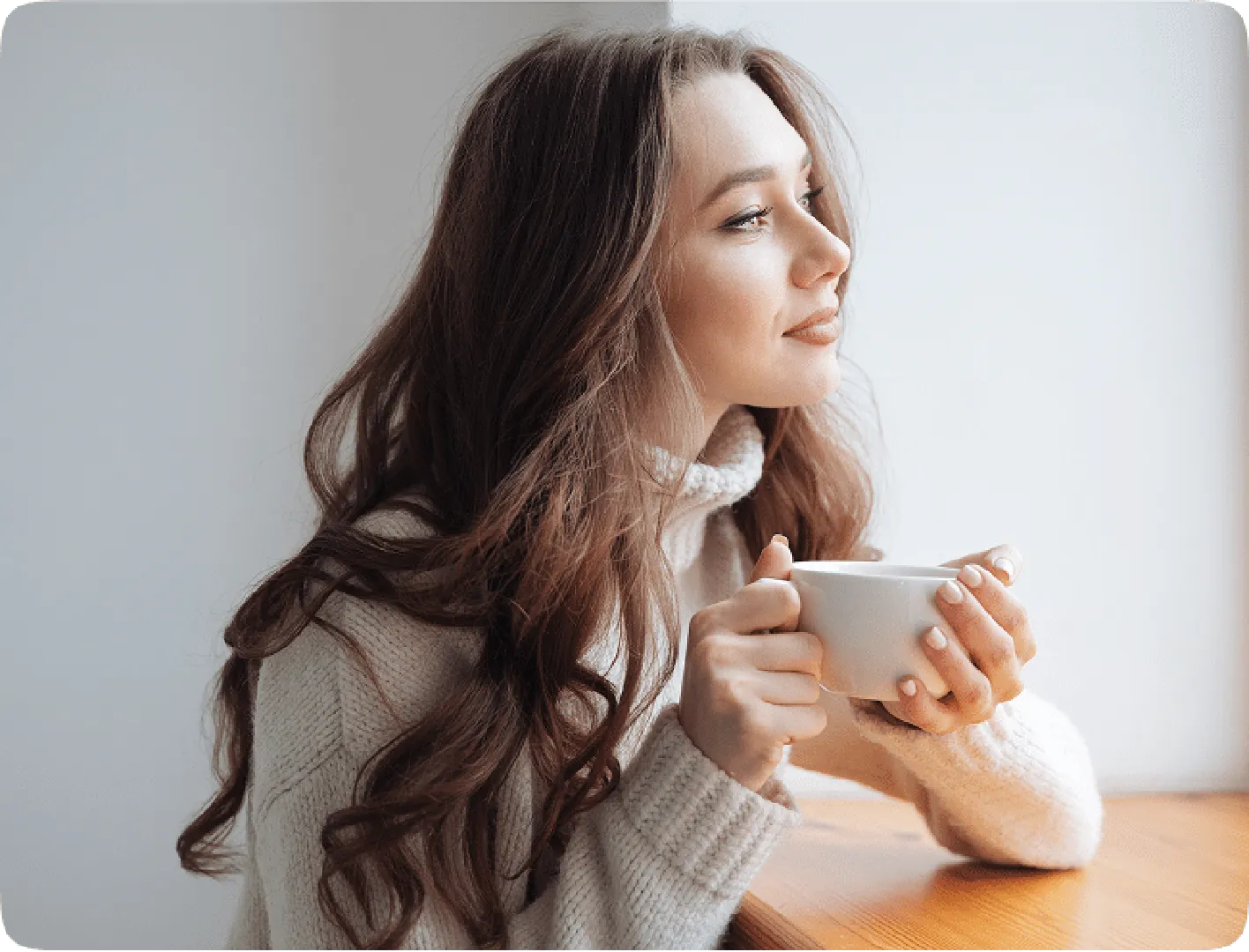 Woman in a white sweater sitting, holding a cup and looking at a distance with a smiley face.
