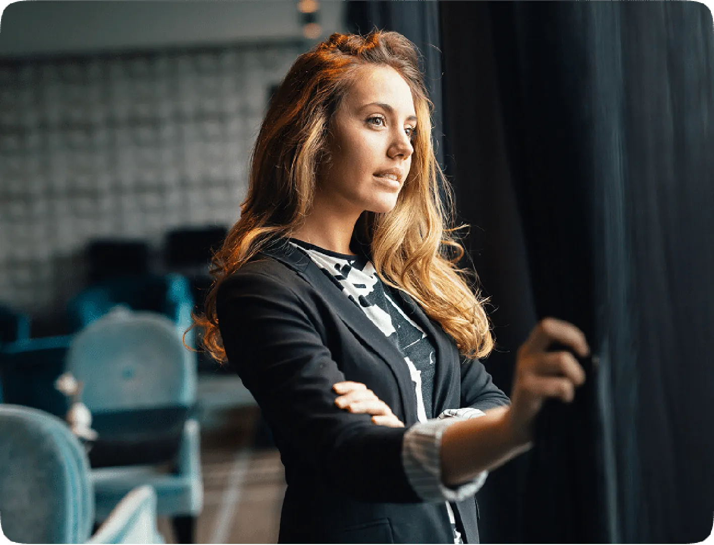 Woman in black blazer by window, gazing thoughtfully as soft natural light highlights her hair.