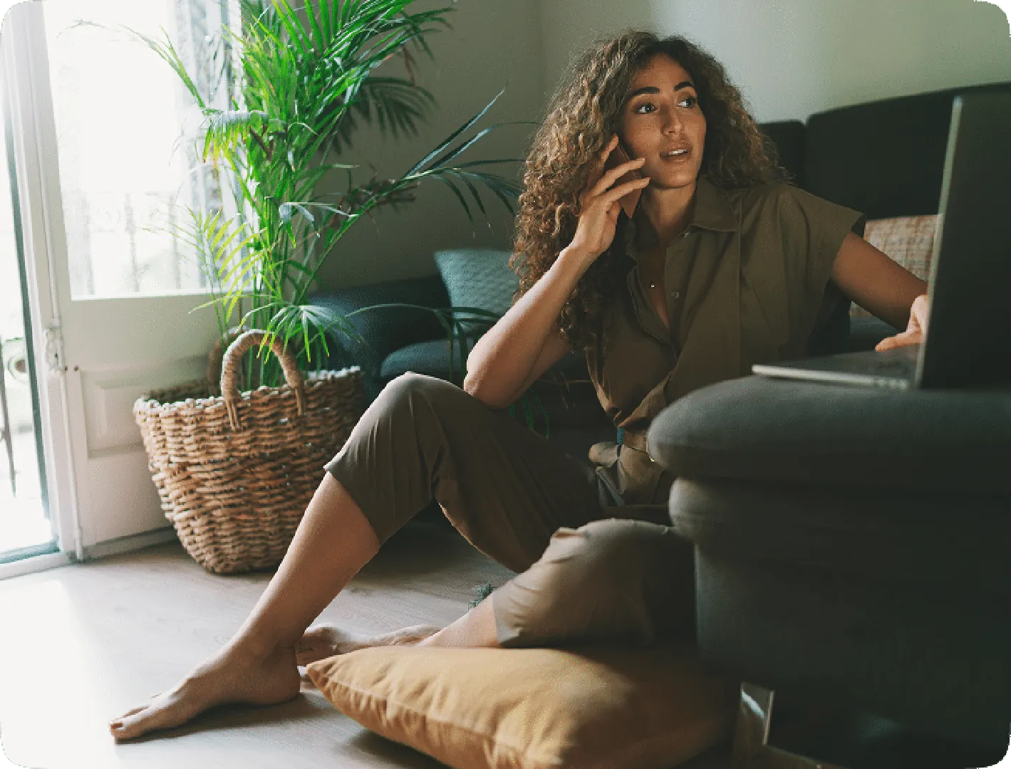 Woman sitting on floor with pillows, using laptop and talking on phone, with green grass behind.