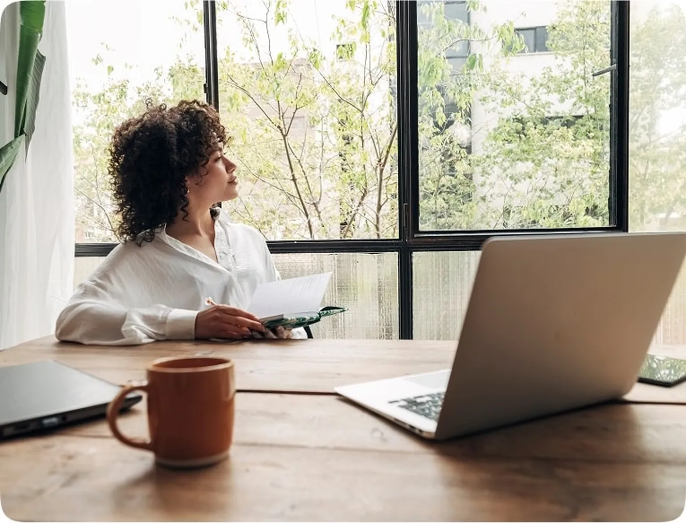 Woman with curly hair in white shirt at table with laptop, holding pen and gazing outside.