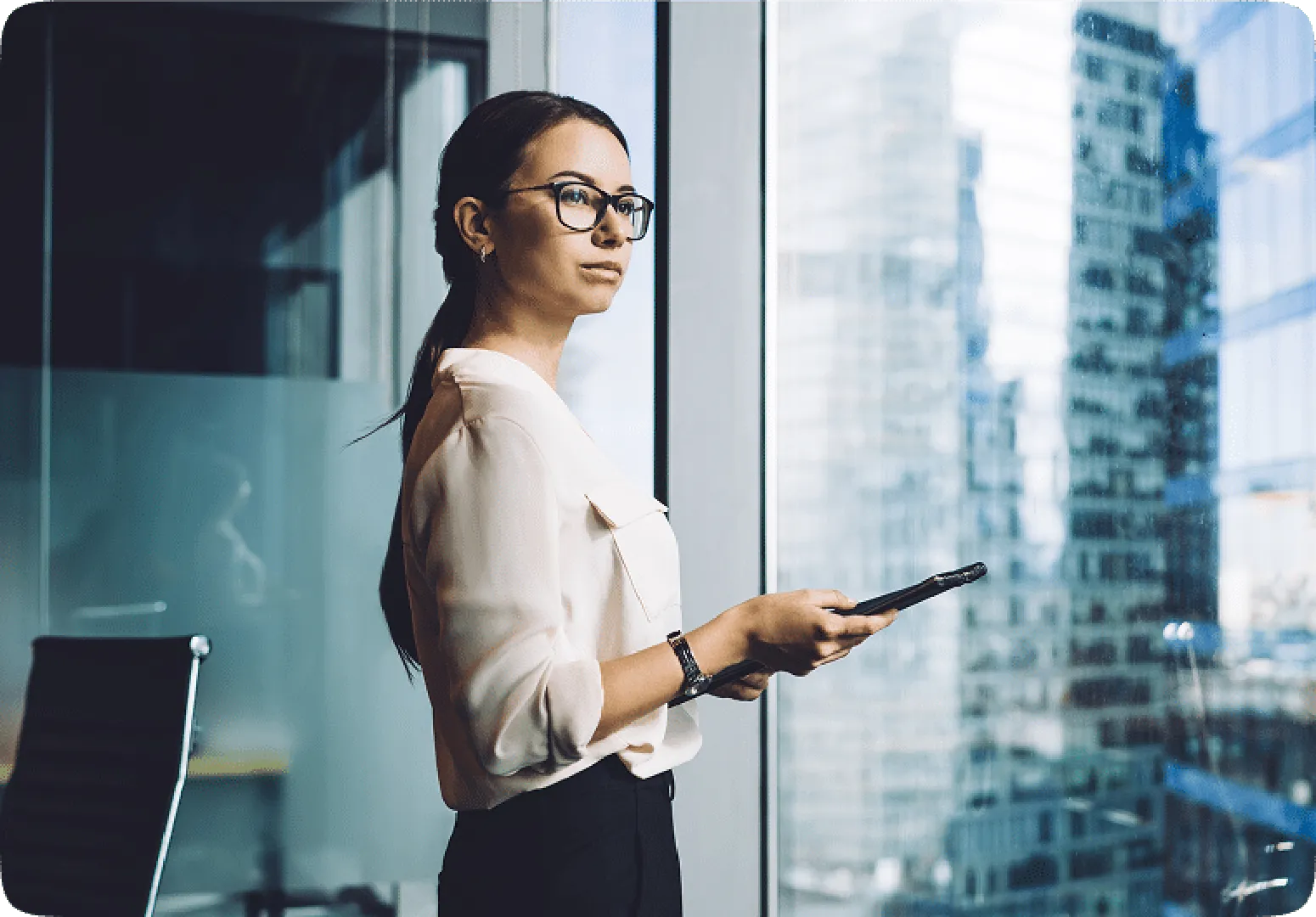 Woman in glass and white dress standing by window holding a device, gazing thoughtfully.
